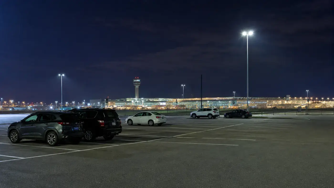 Parked cars waiting in a lit airport waiting lot at night with the terminal illuminated in the distance