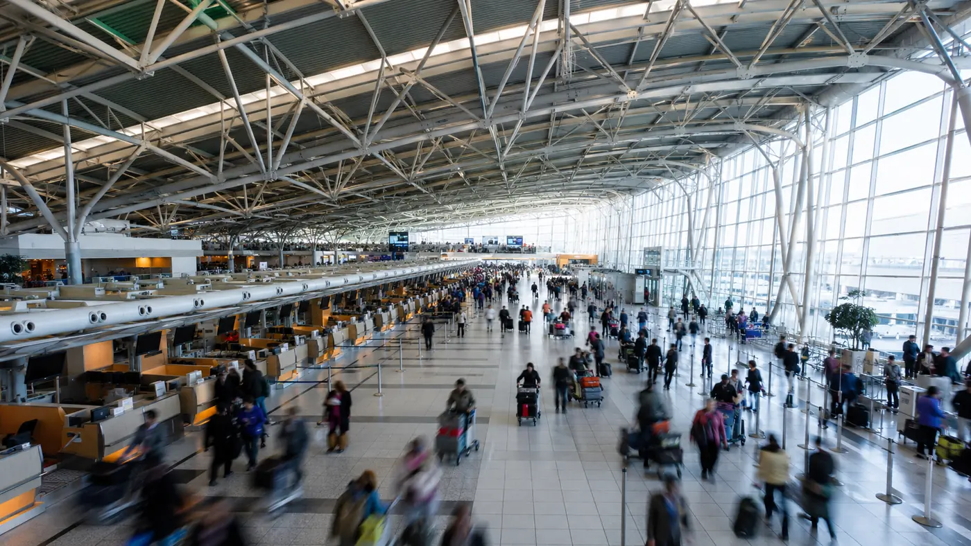 Wide check-in hall with long rows of counters and travellers with luggage trolleys under a high glass roof