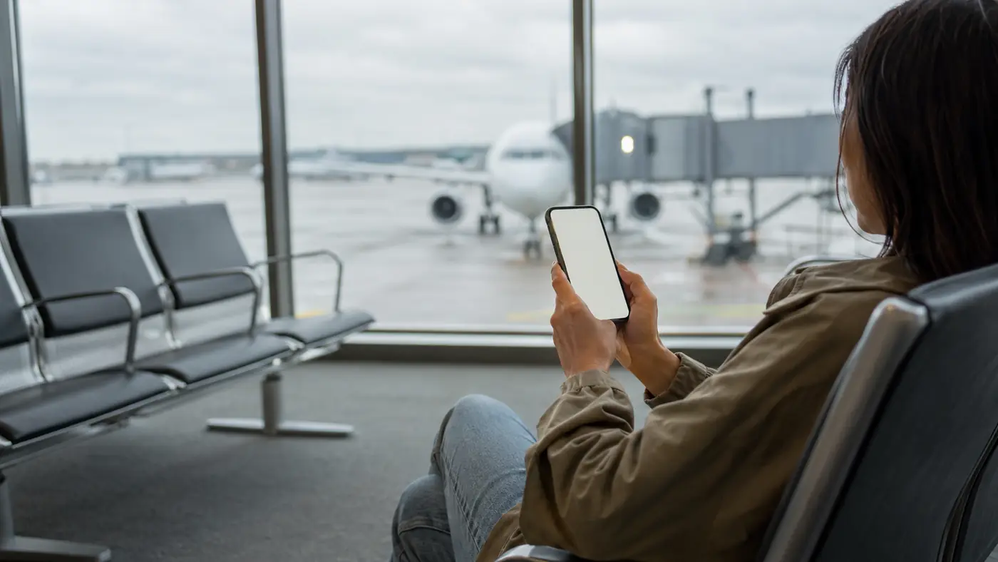 Person seated in an airport gate area holding a phone, with an aircraft visible through the window behind